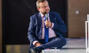 Adrian Klarenbach w garniturze, z mikrofonem w dłoni, siedzi na scenie. Mówi o swojej chorobie.