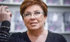Aleksandra Jakubowska obecnie, w okularach, z dłonią przy włosach, na tle półek z książkami.