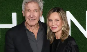 Harrison Ford i jego żona Calista Flockhart uśmiechają się do aparatu na tle zielonej ściany z białymi literami.