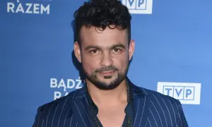Robert El Gendy w granatowej marynarce w paski, z charakterystyczną fryzurą i zarostem, pozuje na tle niebieskiego banera z logo TVP.