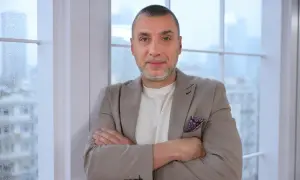 Maciej Dębosz prywatnie, w beżowej marynarce i z dumnym spojrzeniem, opiera się o okno.