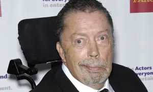 Tim Curry w wózku inwalidzkim, zmagający się z chorobą, ale wciąż z iskrą w oku.