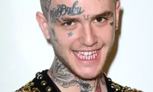Uśmiechnięty Lil Peep z tatuażami na twarzy i szyi, błyszczącymi różowymi zębami. Pytanie czy lil peep żyje, pozostaje w sercach fanów.