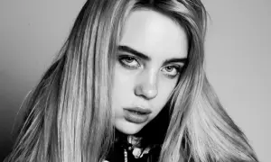 Zbliżenie na twarz Billie Eilish, artystki znanej z unikalnych utworów. Jej spojrzenie jest intensywne, a długie włosy opadają na ramiona.