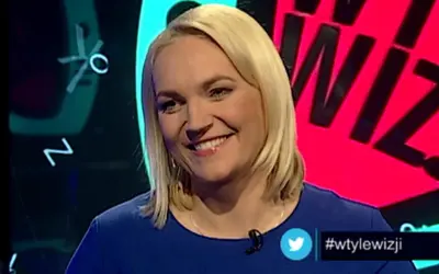 Uśmiechnięta blondynka w niebieskiej sukience. W tle wizji prowadzący, litery i cyfry.