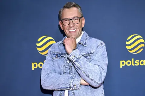 Uśmiechnięty Darek Stolarz w dżinsowej kurtce, z podpartą brodą, na tle logo Polsatu.