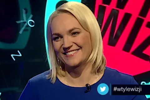 Uśmiechnięta blondynka w niebieskiej sukience. W tle wizji prowadzący, litery i cyfry.