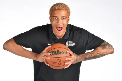 Młody polak w NBA, z blond włosami i tatuażami na ramionach, trzyma piłkę do koszykówki.