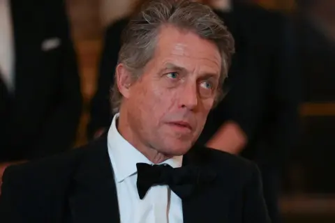 Hugh Grant w czarnym smokingu i muszce, z siwiejącymi włosami, wygląda na zamyślonego.