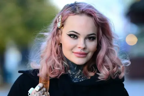 Monika Miller z różowymi włosami i tatuażami na szyi, mimo choroby, uśmiecha się do obiektywu.