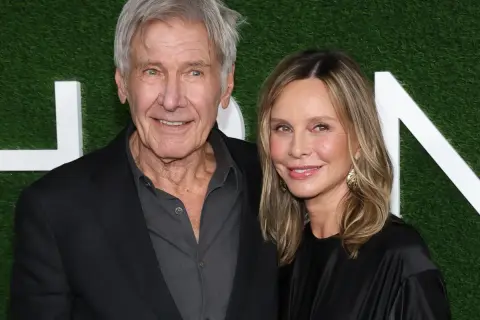 Harrison Ford i jego żona Calista Flockhart uśmiechają się do aparatu na tle zielonej ściany z białymi literami.