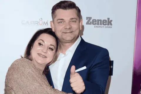 Danuta Martyniuk, w złotej sukience, przytula się do mężczyzny w granatowym garniturze, który pokazuje kciuk w górę.
