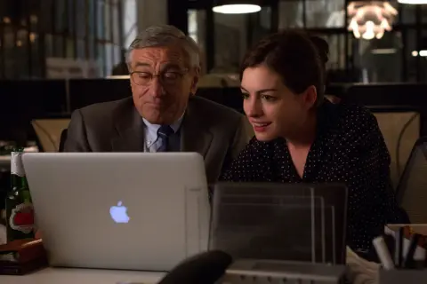 Robert De Niro i Anne Hathaway pracują przy laptopie. Młoda praktykantka z entuzjazmem analizuje dane, podczas gdy doświadczony szef czuwa nad jej pracą.