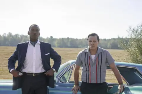 Obsada filmu Green Book: Mahershala Ali w eleganckim garniturze i Viggo Mortensen w koszuli z lat 60. stoją obok siebie przy samochodzie.