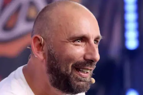 Radosław Bielecki, uśmiechnięty, z zarostem, w białej koszulce. Mimo choroby, widać w nim siłę i determinację.