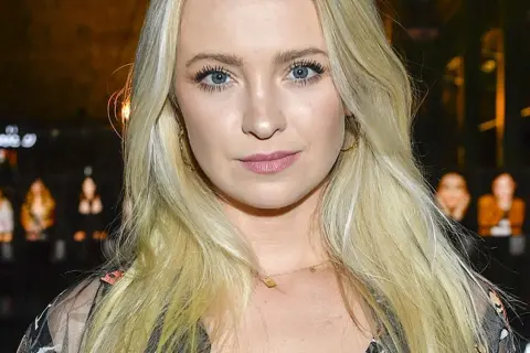 Blondynka z niebieskimi oczami, w złotych kolczykach, wygląda jakby była w centrum uwagi, może nawet w związku z jakąś aferą, jak kurdej szatan afera.