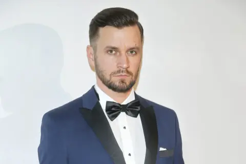 Piotr Domalewski w granatowym garniturze i czarnej muszce. Wygląda jakby właśnie wracał do żony po ważnym wydarzeniu.