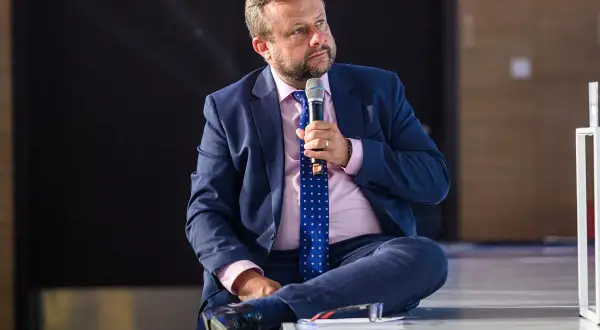 Adrian Klarenbach w garniturze, z mikrofonem w dłoni, siedzi na scenie. Mówi o swojej chorobie.
