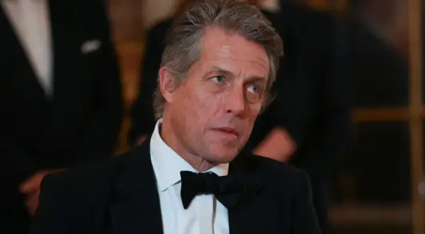 Hugh Grant w czarnym smokingu i muszce, z siwiejącymi włosami, wygląda na zamyślonego.