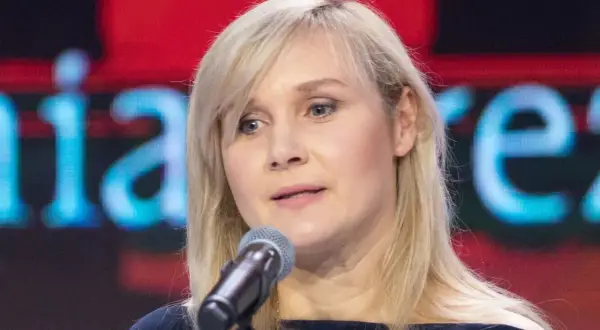 Kobieta z blond włosami mówi do mikrofonu na tle ekranu z napisem.