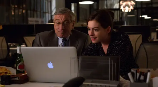Robert De Niro i Anne Hathaway pracują przy laptopie. Młoda praktykantka z entuzjazmem analizuje dane, podczas gdy doświadczony szef czuwa nad jej pracą.