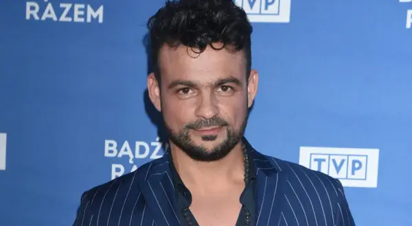 Robert El Gendy w granatowej marynarce w paski, z charakterystyczną fryzurą i zarostem, pozuje na tle niebieskiego banera z logo TVP.