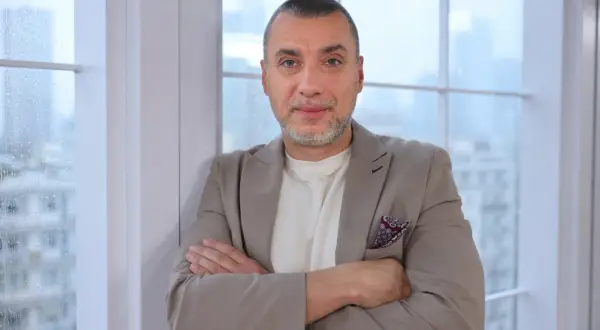 Maciej Dębosz prywatnie, w beżowej marynarce i z dumnym spojrzeniem, opiera się o okno.