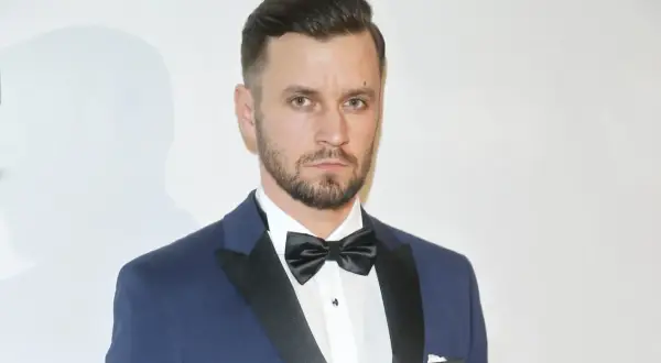 Piotr Domalewski w granatowym garniturze i czarnej muszce. Wygląda jakby właśnie wracał do żony po ważnym wydarzeniu.