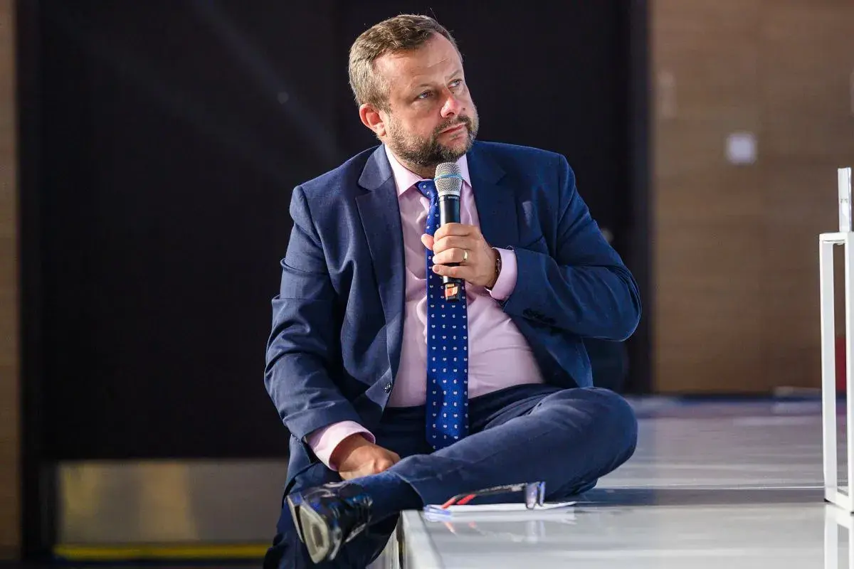 Adrian Klarenbach w garniturze, z mikrofonem w dłoni, siedzi na scenie. Mówi o swojej chorobie.