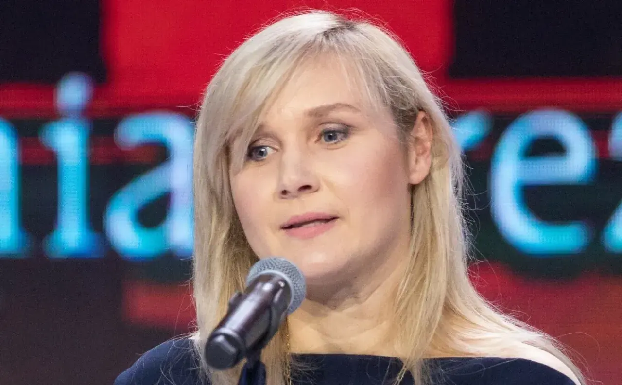 Kobieta z blond włosami mówi do mikrofonu na tle ekranu z napisem.