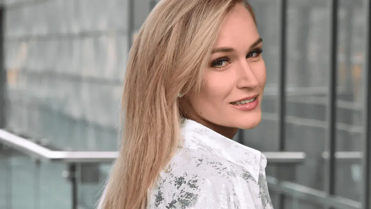 Uśmiechnięta Kamila Boś, w białej koszuli z wzorem, patrzy przez ramię.