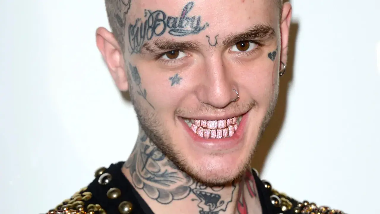 Uśmiechnięty Lil Peep z tatuażami na twarzy i szyi, błyszczącymi różowymi zębami. Pytanie czy lil peep żyje, pozostaje w sercach fanów.