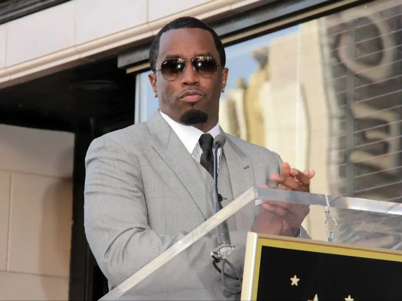 P Diddy w szarym garniturze i okularach przeciwsłonecznych przemawia przy mównicy.