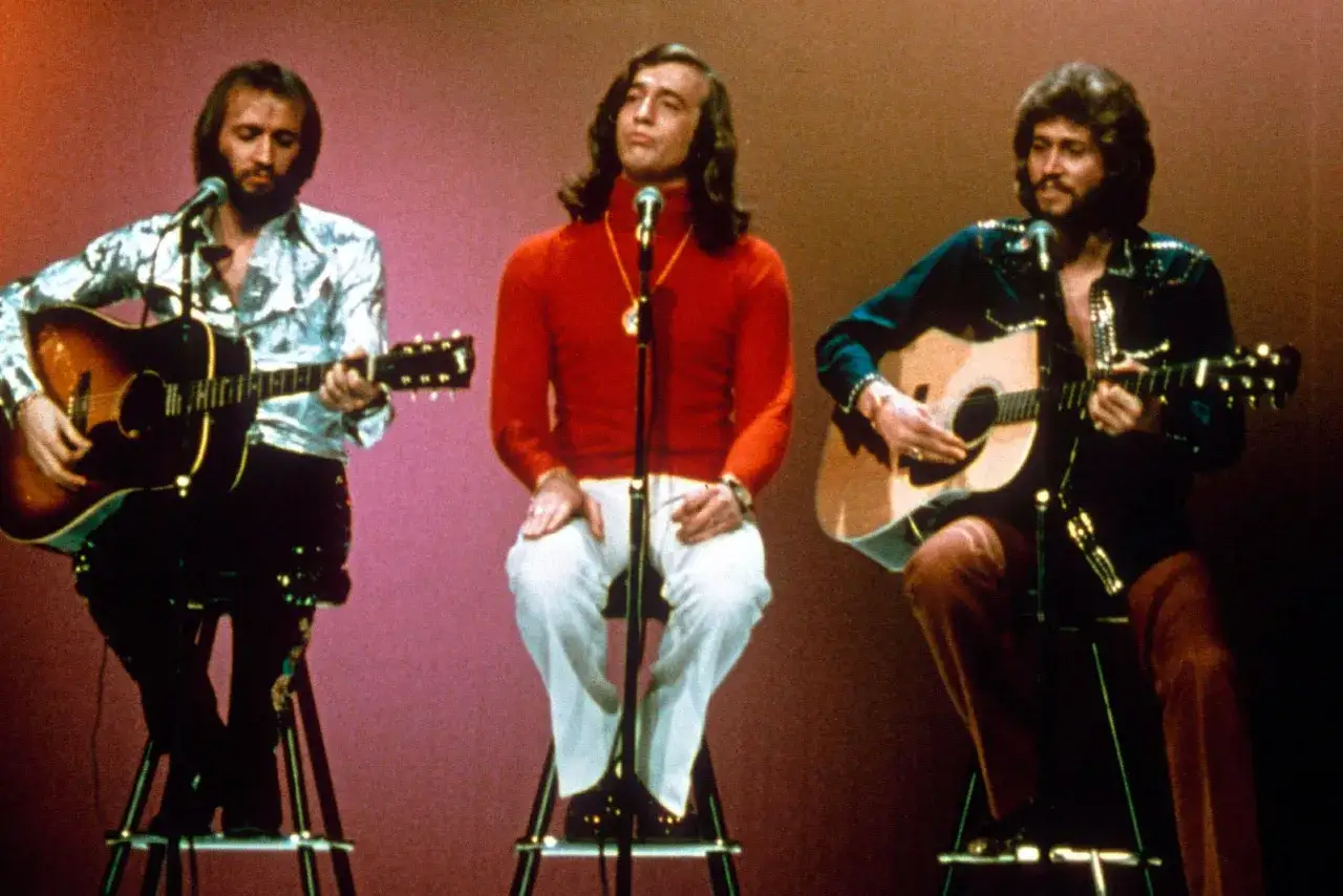 Trzej bracia z Bee Gees, Barry, Robin i Maurice Gibb, siedzą na scenie z gitarami. Kto żyje? Wszyscy trzej!