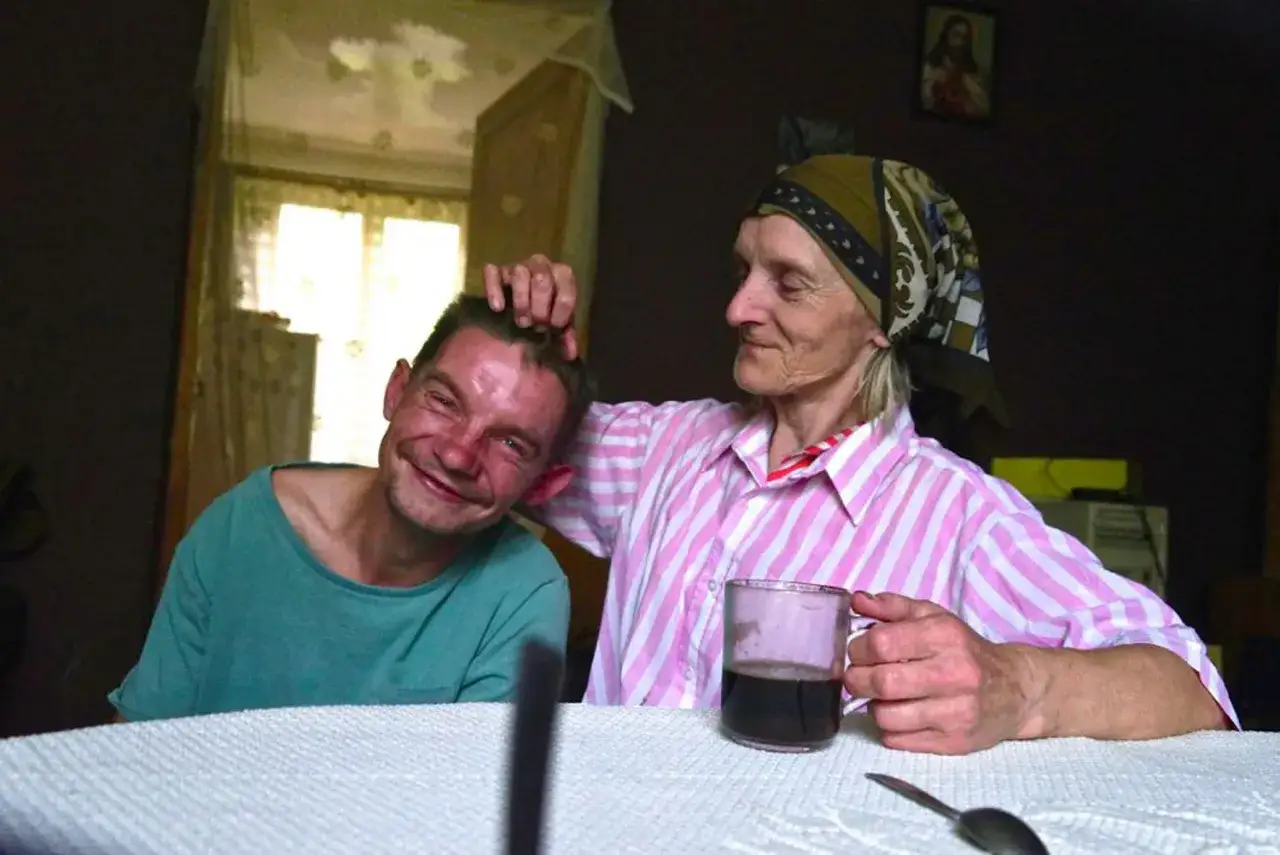 Uśmiechnięty mężczyzna z bandziorkiem na twarzy, którego głaszcze po głowie starsza kobieta w chustce i różowej koszuli.