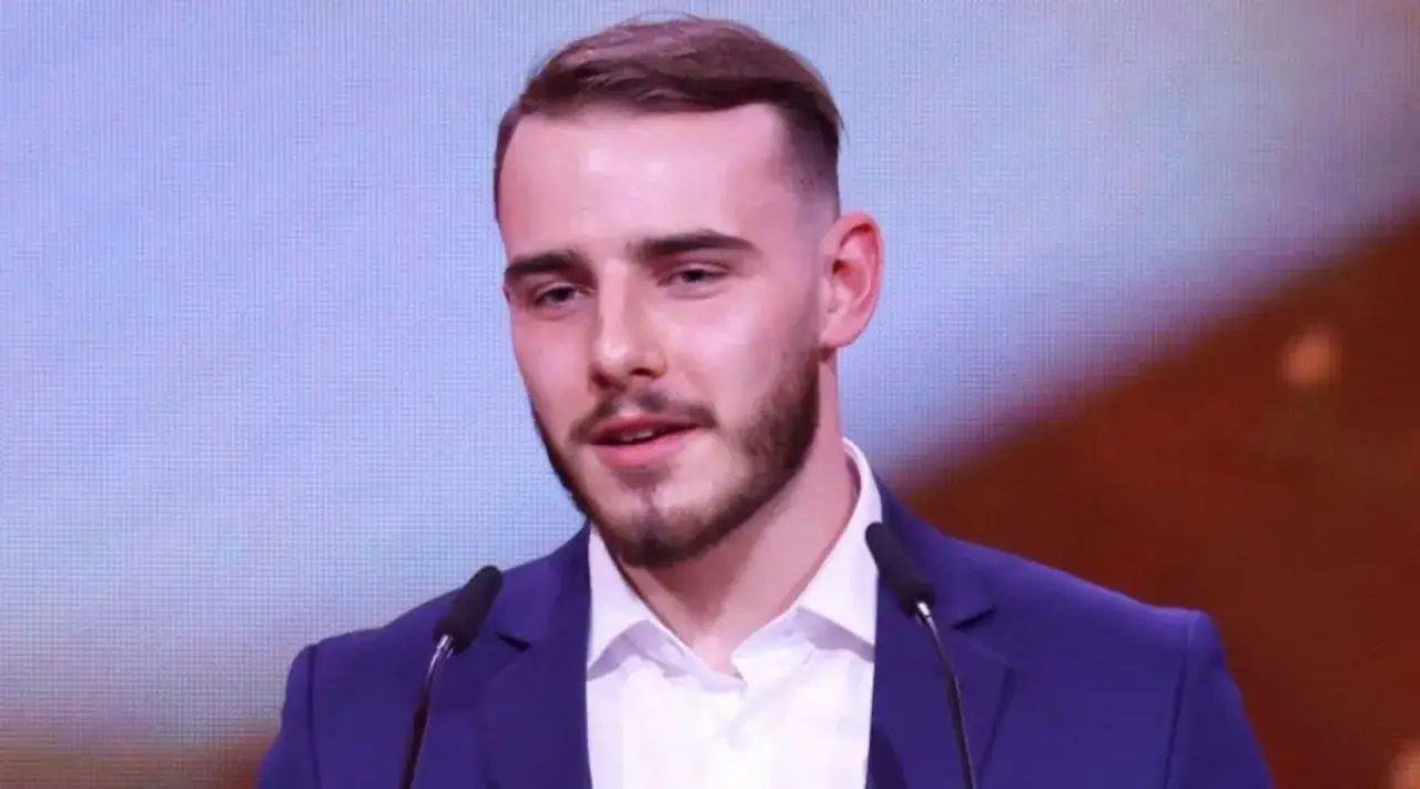 Krystian Ochman w granatowej marynarce, z zarostem, przemawia przy mikrofonie. Może opowiada o swojej żonie.