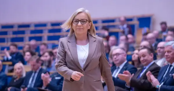 Kobieta w okularach i brązowej marynarce idzie na tle publiczności.