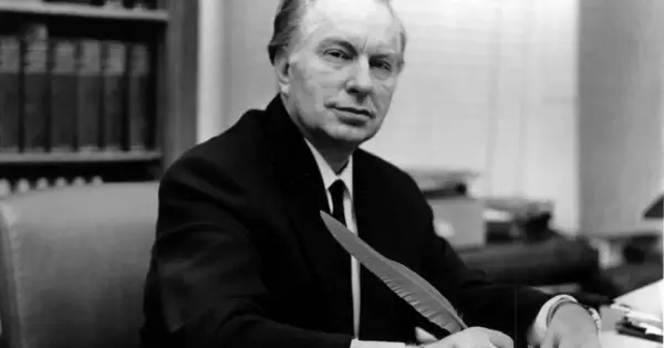 L. Ron Hubbard, twórca scjentologii, siedzi przy biurku z piórem w dłoni, gotów do pisania.