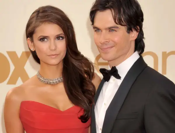 Nina Dobrev w czerwonej sukni i jej partner w smokingu, uśmiechnięci na ściance.