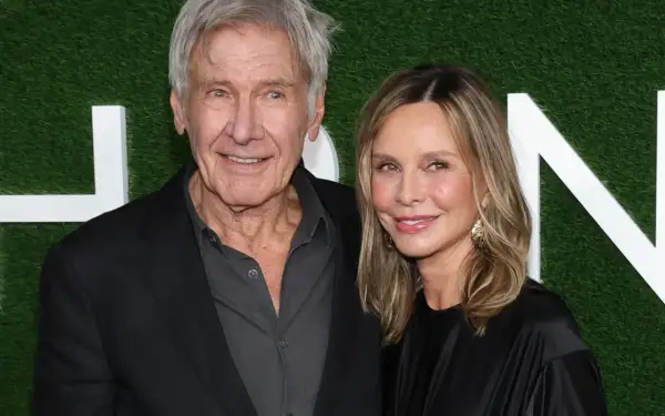 Harrison Ford i jego żona Calista Flockhart uśmiechają się do aparatu na tle zielonej ściany z białymi literami.