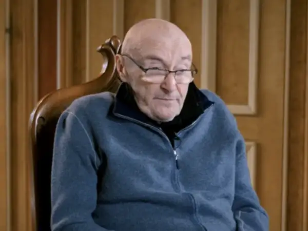 Phil Collins, zmagający się z chorobą, siedzi w fotelu, ubrany w niebieski polar.