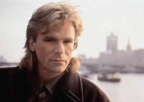 MacGyver obsada: Richard Dean Anderson w płaszczu, z charakterystyczną fryzurą, na tle zamglonego miasta.