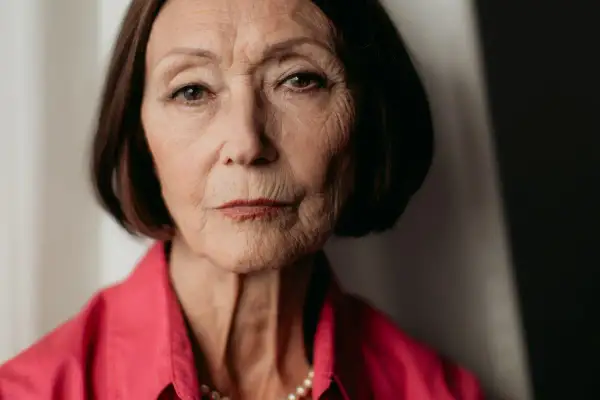 Portret starszej kobiety, Ewy Woydyłło, z widocznymi zmarszczkami, w różowej bluzce i perłach. Jej życie prywatne jest pełne doświadczeń.
