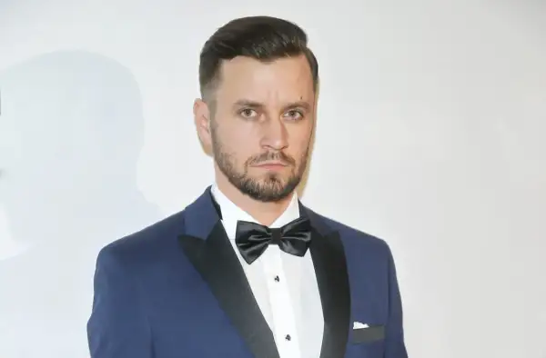 Piotr Domalewski w granatowym garniturze i czarnej muszce. Wygląda jakby właśnie wracał do żony po ważnym wydarzeniu.